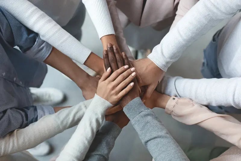 A group of people stand in a circle with their hands stacked together in a gesture of unity and teamwork.